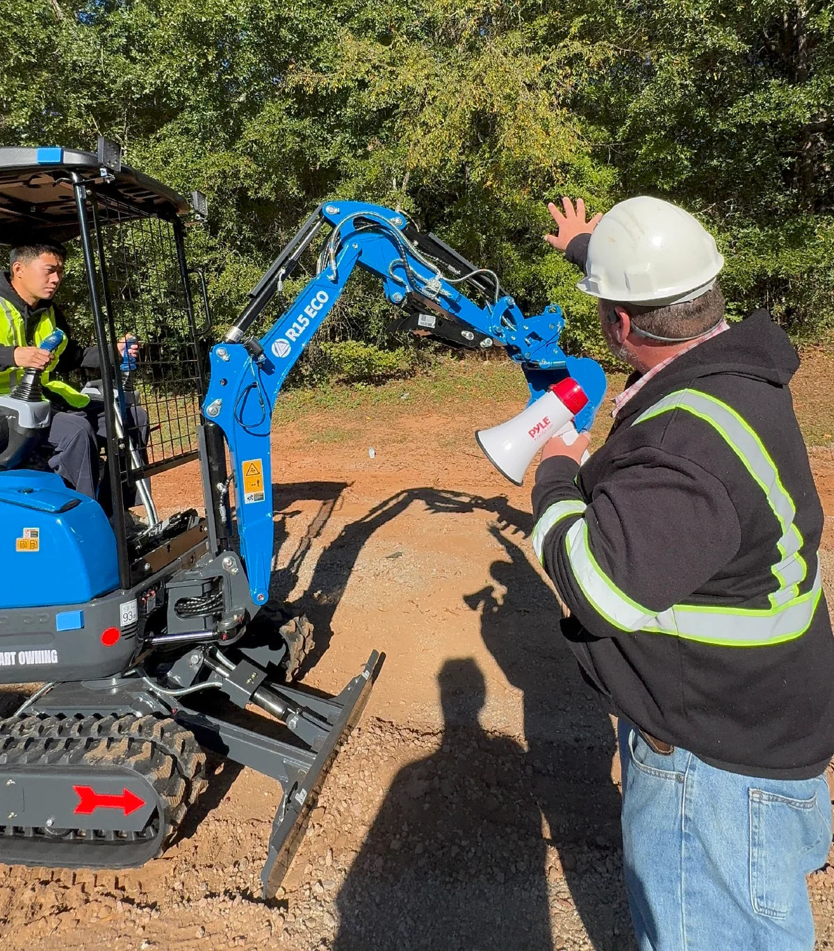 Instructor guiding an operator in a blue mini excavator during an outdoor GHM class